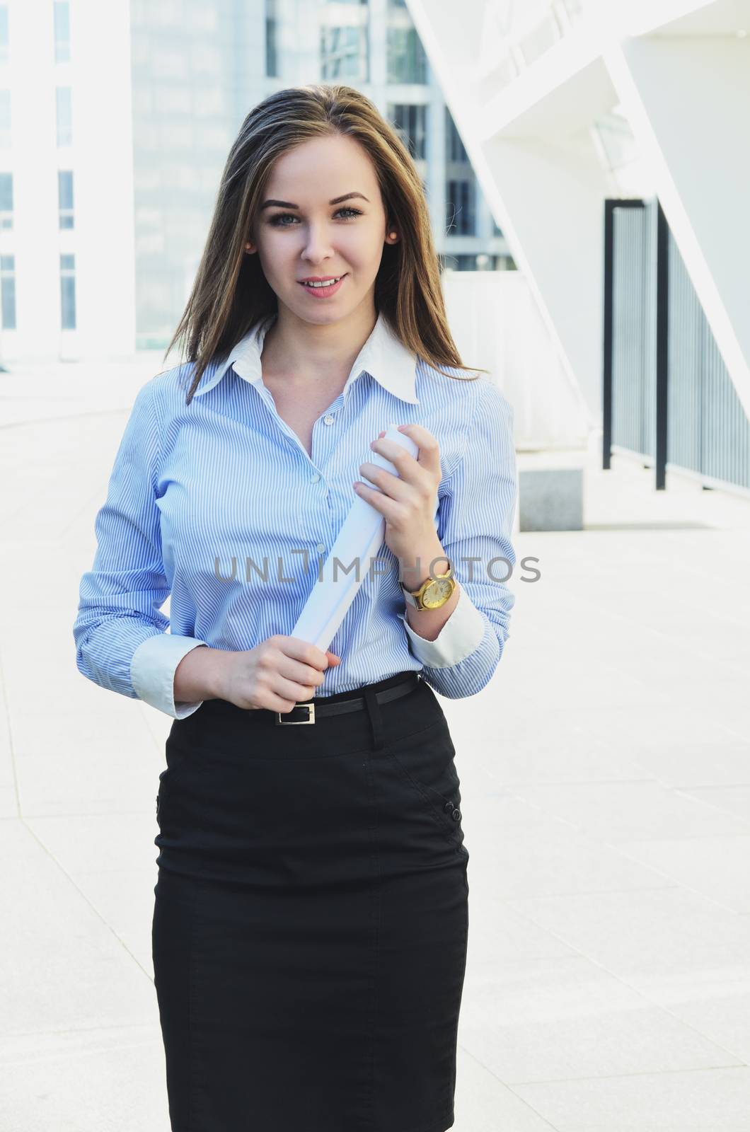 Young attractive engineer girl looks into the camera holding a roll of documents, being near a beautiful tall building