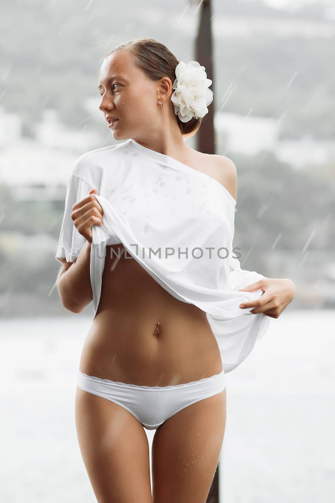 Attractive young female in while blouse and panties embracing herself and keeping eyes closed while standing near water on windy day