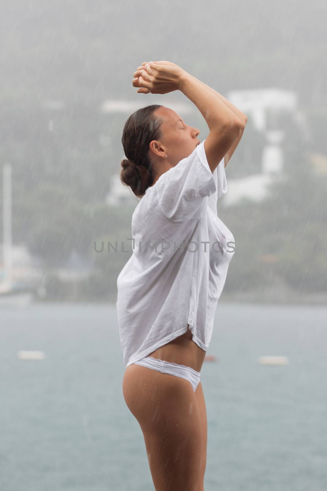 Attractive young female in while blouse and panties embracing herself and keeping eyes closed while standing near water on windy day