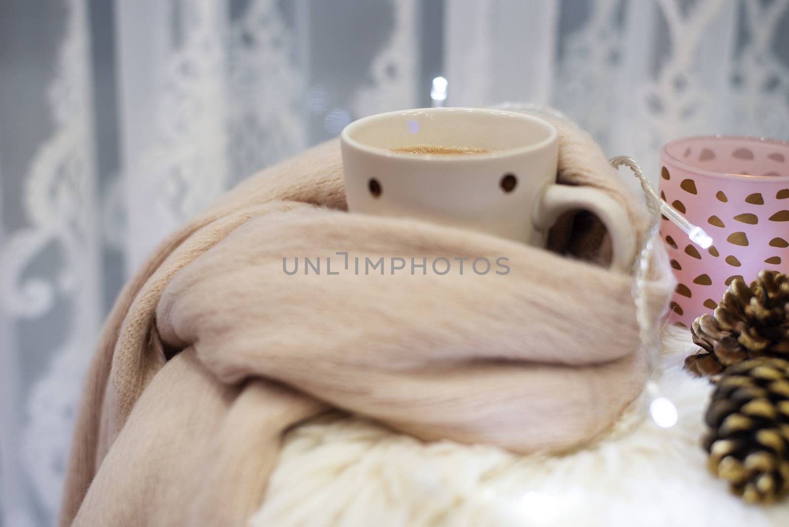 Hot chocolate, a cup of cappuccino on a fur chair in front of a large window with a white sheers curtain. Warm scarf, cones and lights around. Cozy winter evenings by sevda_stancheva