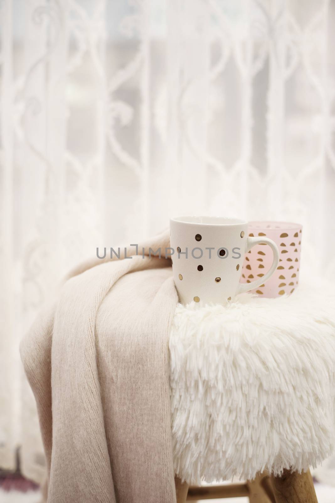 Hot chocolate, a cup of cappuccino on a fur chair in front of a large window with a white sheers curtain. Warm scarf and lights around. Cozy winter eveningsHot chocolate, a cup of cappuccino on a fur chair in front of a large window with a white sheers curtain. Warm scarf and lights around. Cozy winter evenings by sevda_stancheva