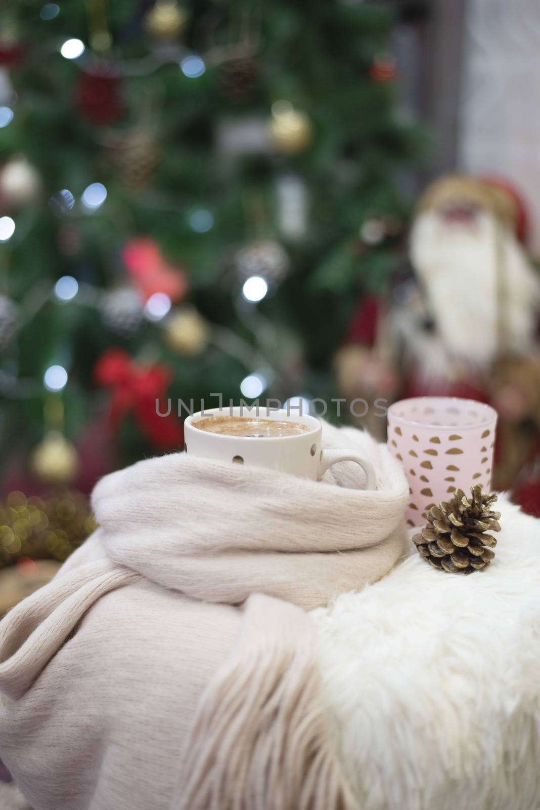 Christmas time. Hot chocolate, a cup of cappuccino on a fur chair in front of a large Christmas tree with balls and lights. Warm scarf, cones around.