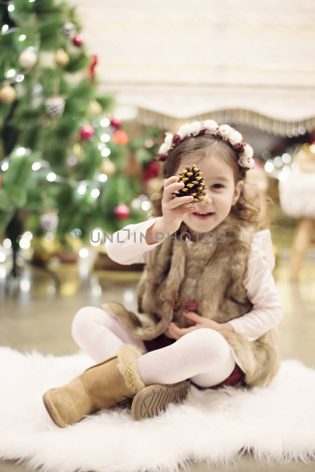 Cute little girl celebrating Christmas at home. She sits on a chair and holds a spruce cone in her hands. Positive emotions. Christmas tree in background. Happy childhood.