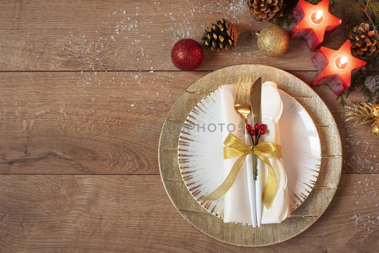 Christmas holiday dinner place setting - plates, napkin, cutlery, gold bauble decorations over oak table background. Fork and spoon on gold plates. Around red candles, cones and balls. Flat lay