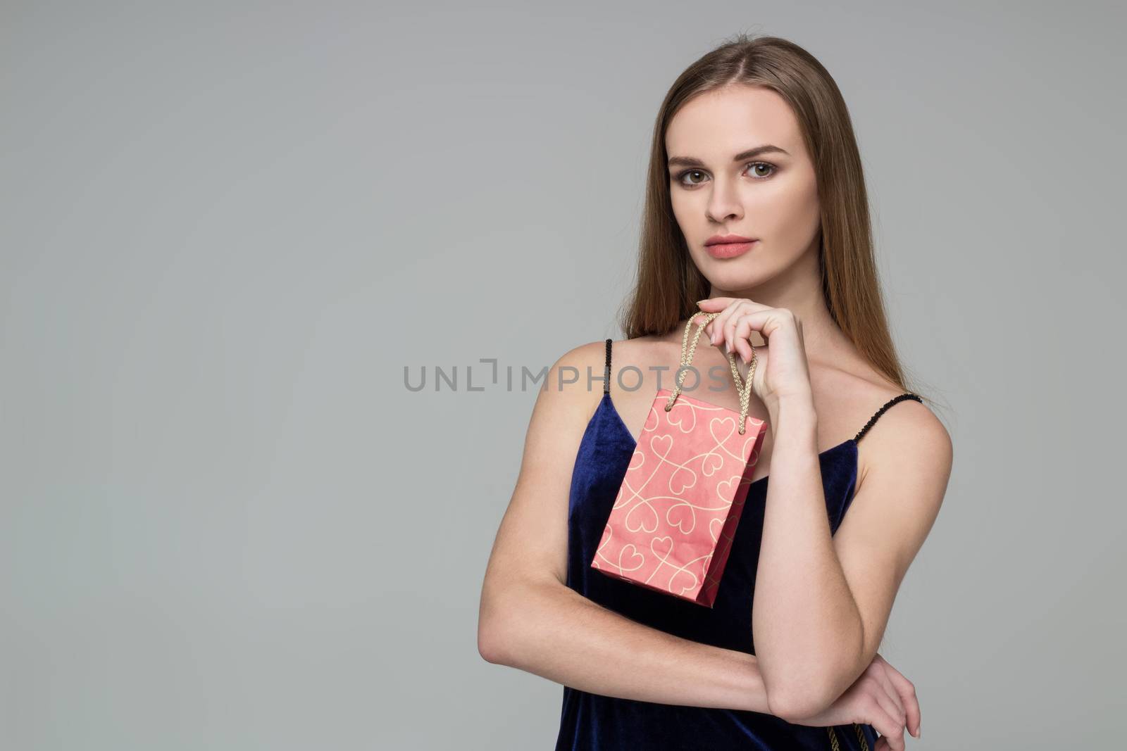 Young model long-haired blond girl in dark blue dress holds red paper gift present package