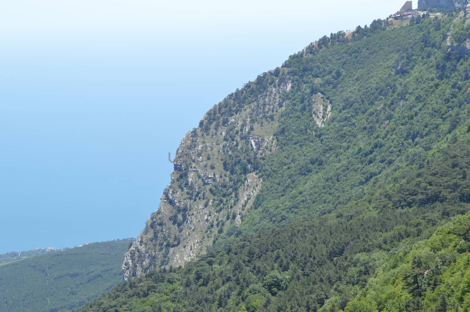 Rocky mountain peaks, steep slopes covered with green forest against blue sky.