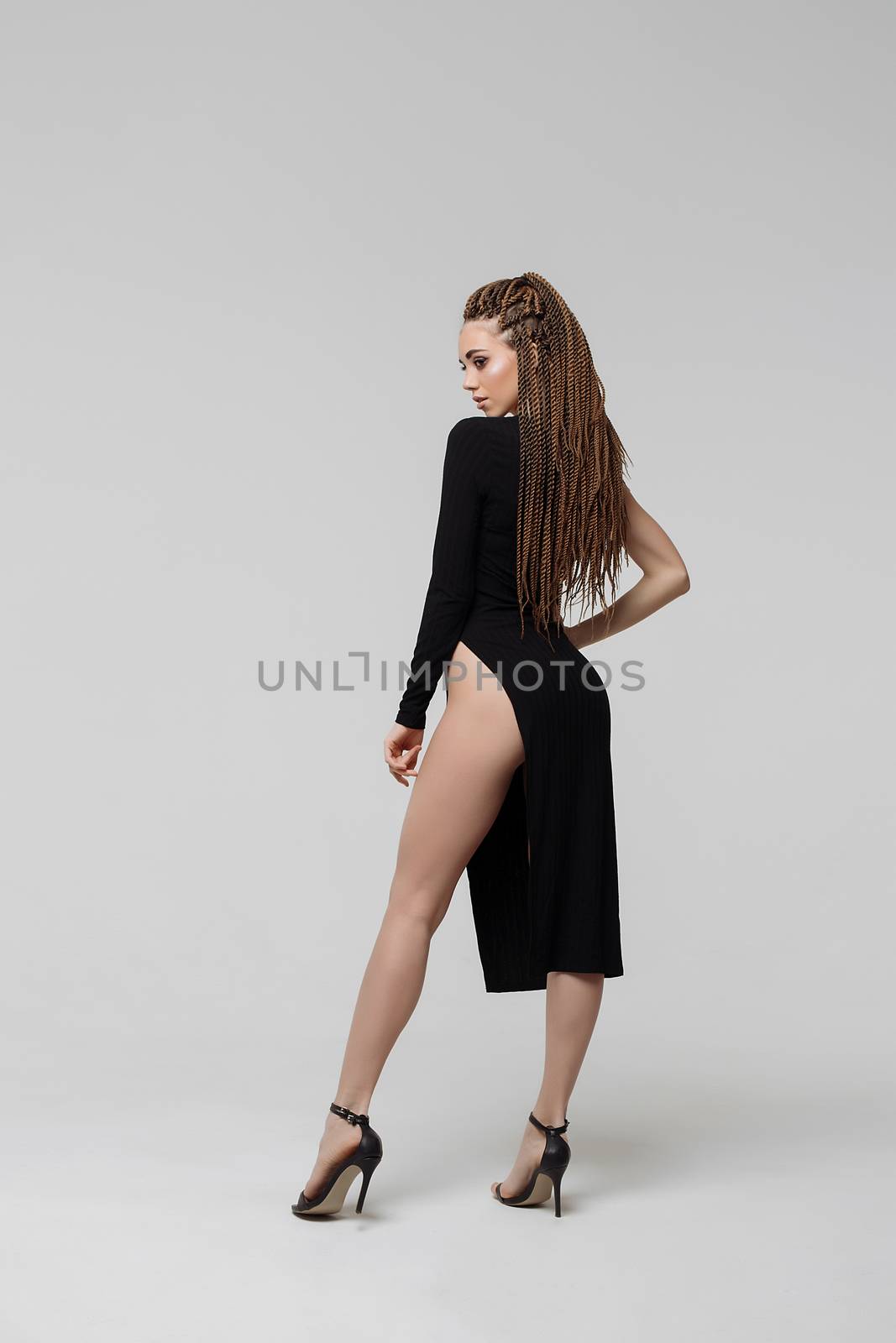 Side view of sensual young female with long dreadlocks wearing amazing black dress and looking at camera while standing on light gray background