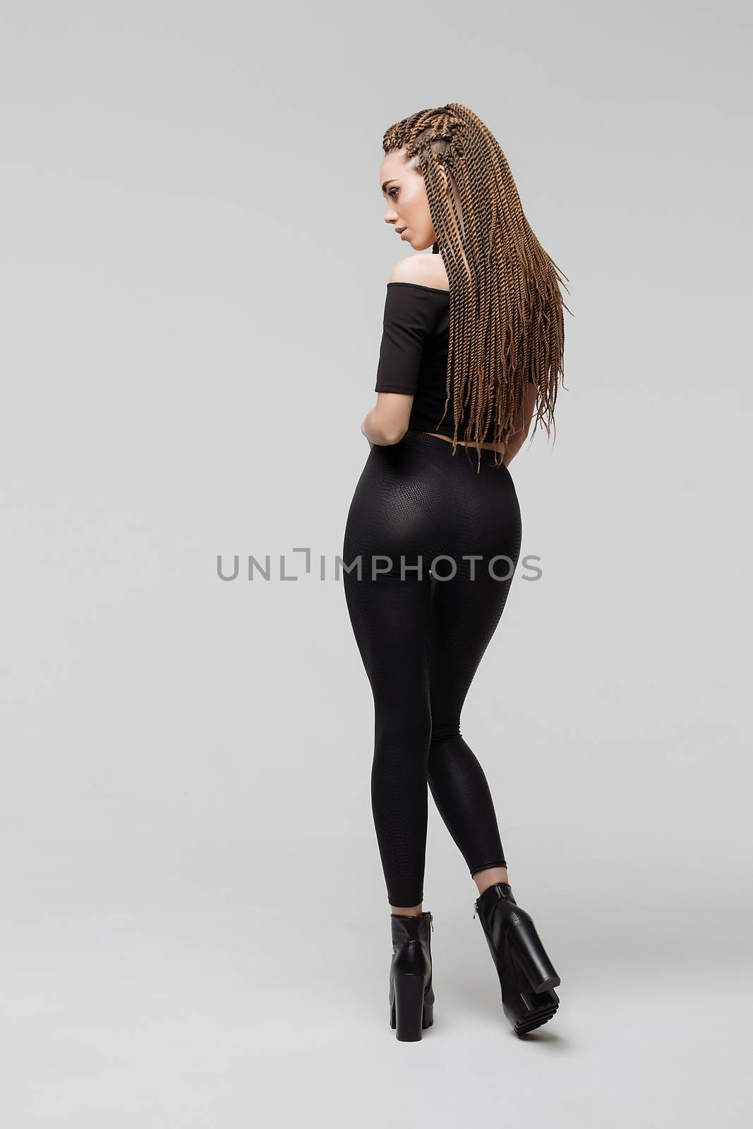 Beautiful young woman with dreadlocks keeping hand on waist and looking at camera while standing on light gray background