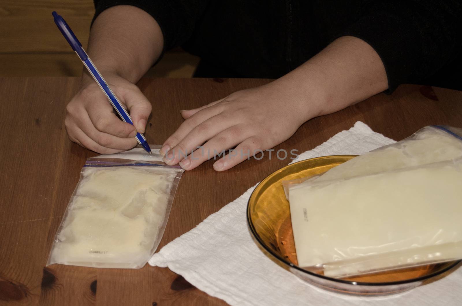 mother packing frozen breast milk in bag for further storage in freezer writing a date when milk was expresed, hands only and wooden table