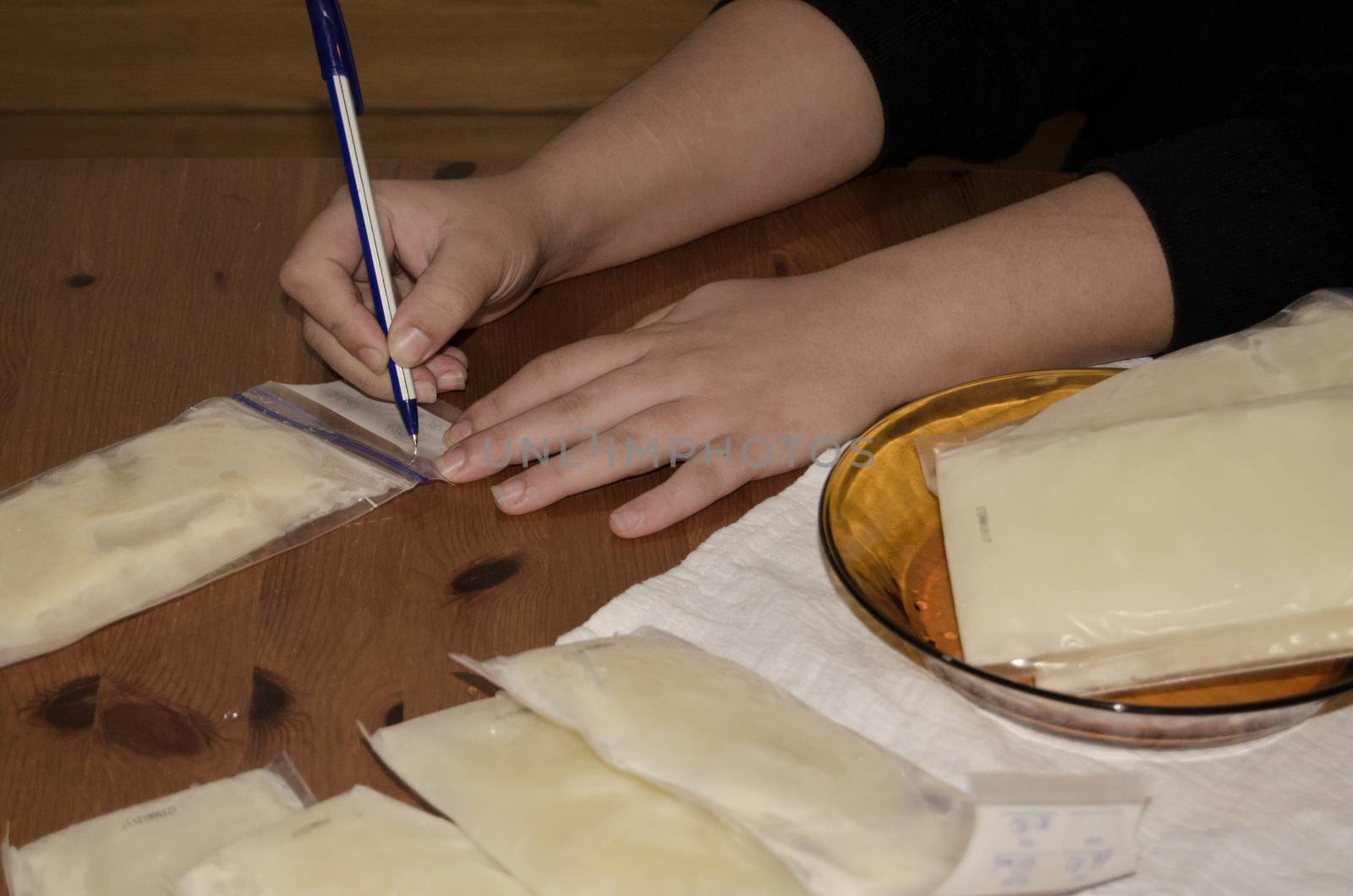 mother packing frozen breast milk in bag for further storage in freezer writing a date when milk was expresed, hands only and wooden table