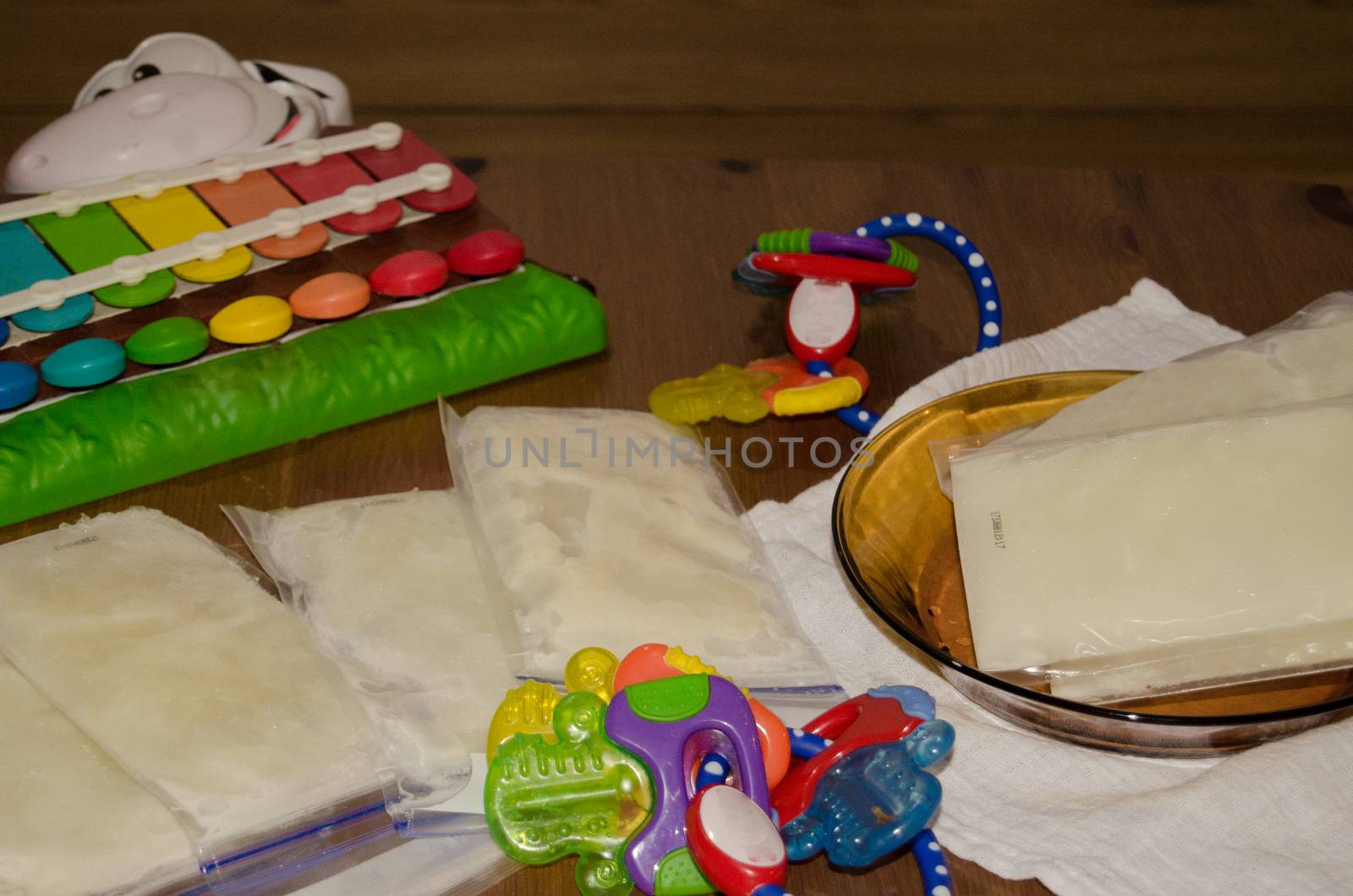 Frozen breast milk in storage bags in glass plate and on the wooden table, surrounded by teething toys and baby cow xylophone, mother milk ready to be stored in the freezer