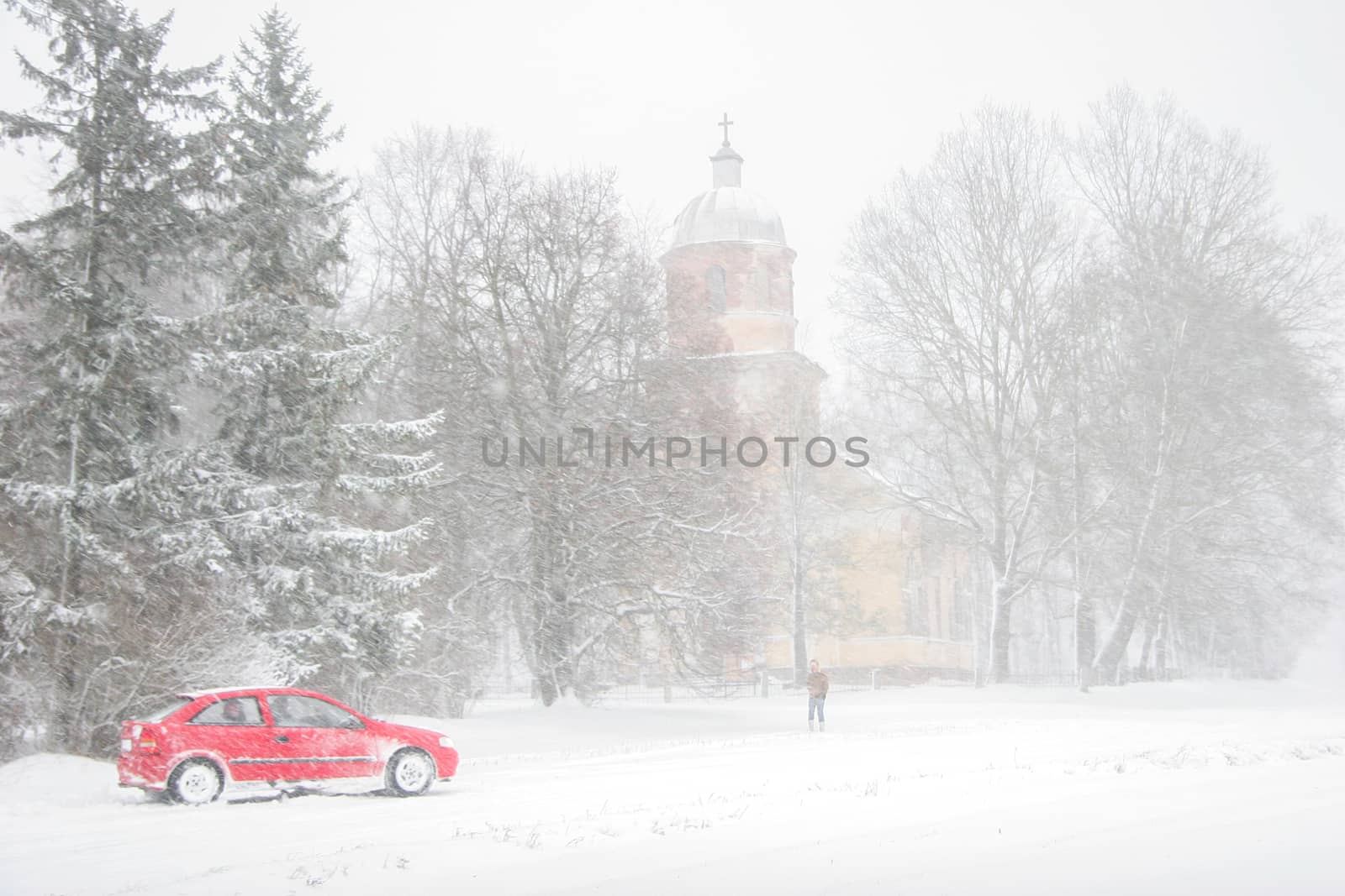 Church in snowfall. Winter in Vecsaule, Latvia. Church covered in snow. Winter landscape with snow covered church, trees and car.



