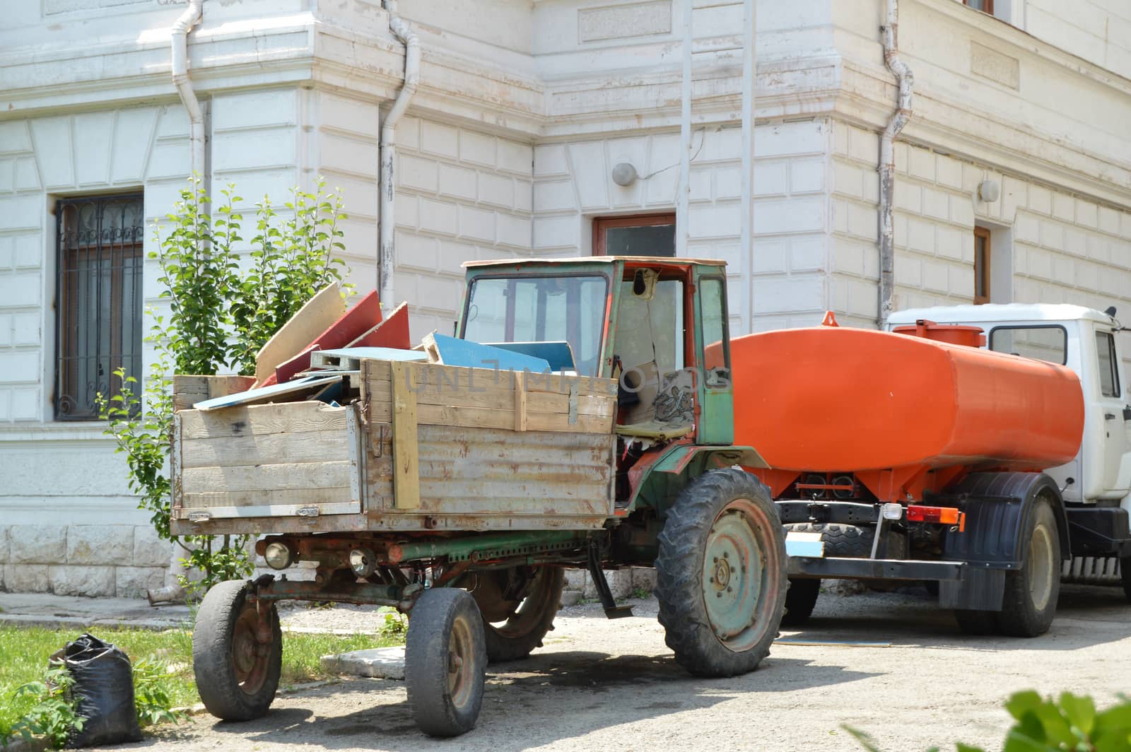 Old tractor with garbage in the back is near the watering machine, cleaning equipment for cleanliness in parks on a Sunny summer day.