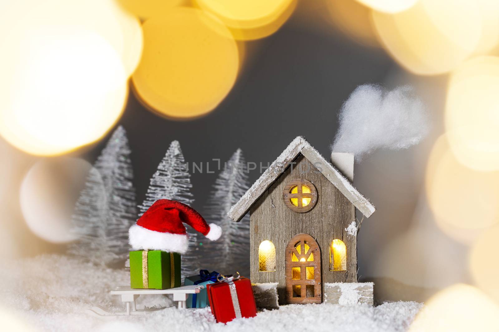 Christmas card with cozy small decorative house , gifts and Santa Claus cap on sleigh on snow over fairy tale winter fir forest background and falling snow . New Year design postcard . Copy space for text