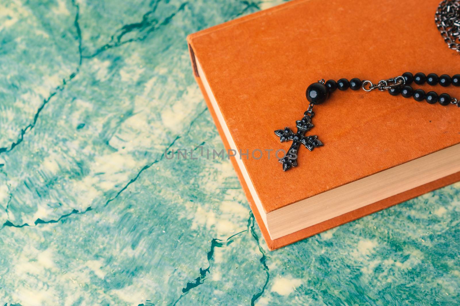 Black rosary and cross resting on the closed book at green table, seen from above.religion school concept.Vintage style.