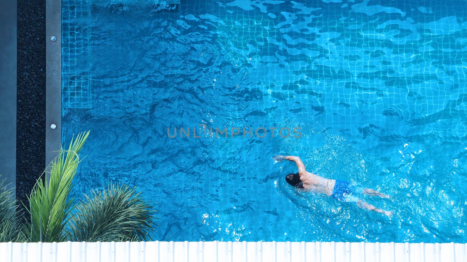 Swimming pool top view angle which young man relaxing and enjoying in pools of his hotel in summer season of Thailand at vacation day and have sun light and flares on blue water surface.