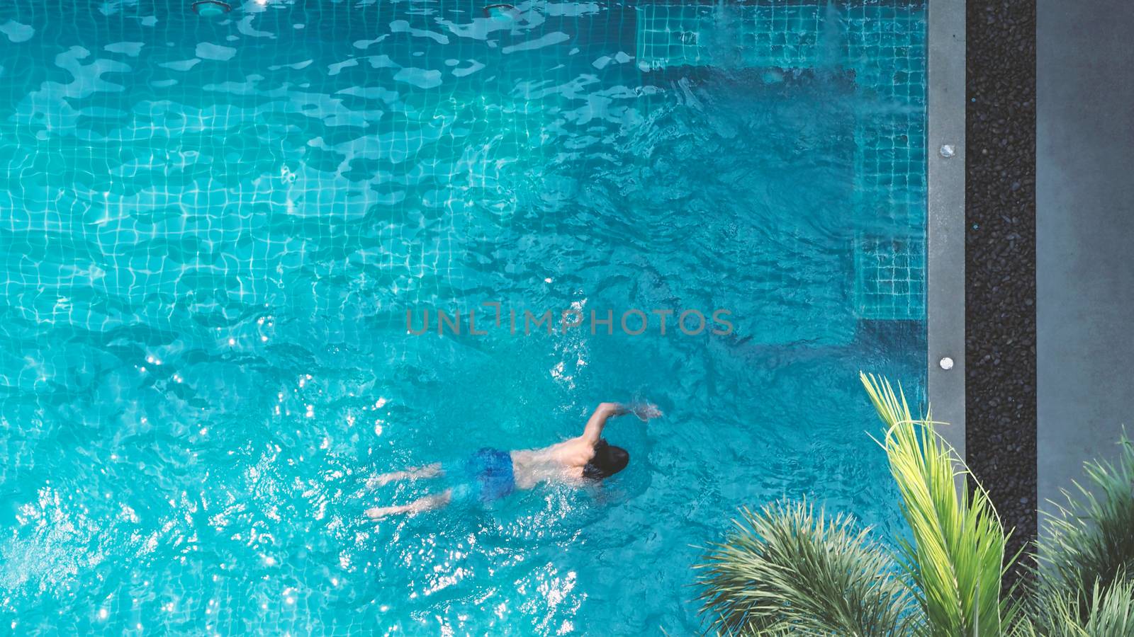 Swimming pool top view angle which young man relaxing and enjoying in pools of his hotel in summer season of Thailand at vacation day and have sun light and flares on blue water surface.