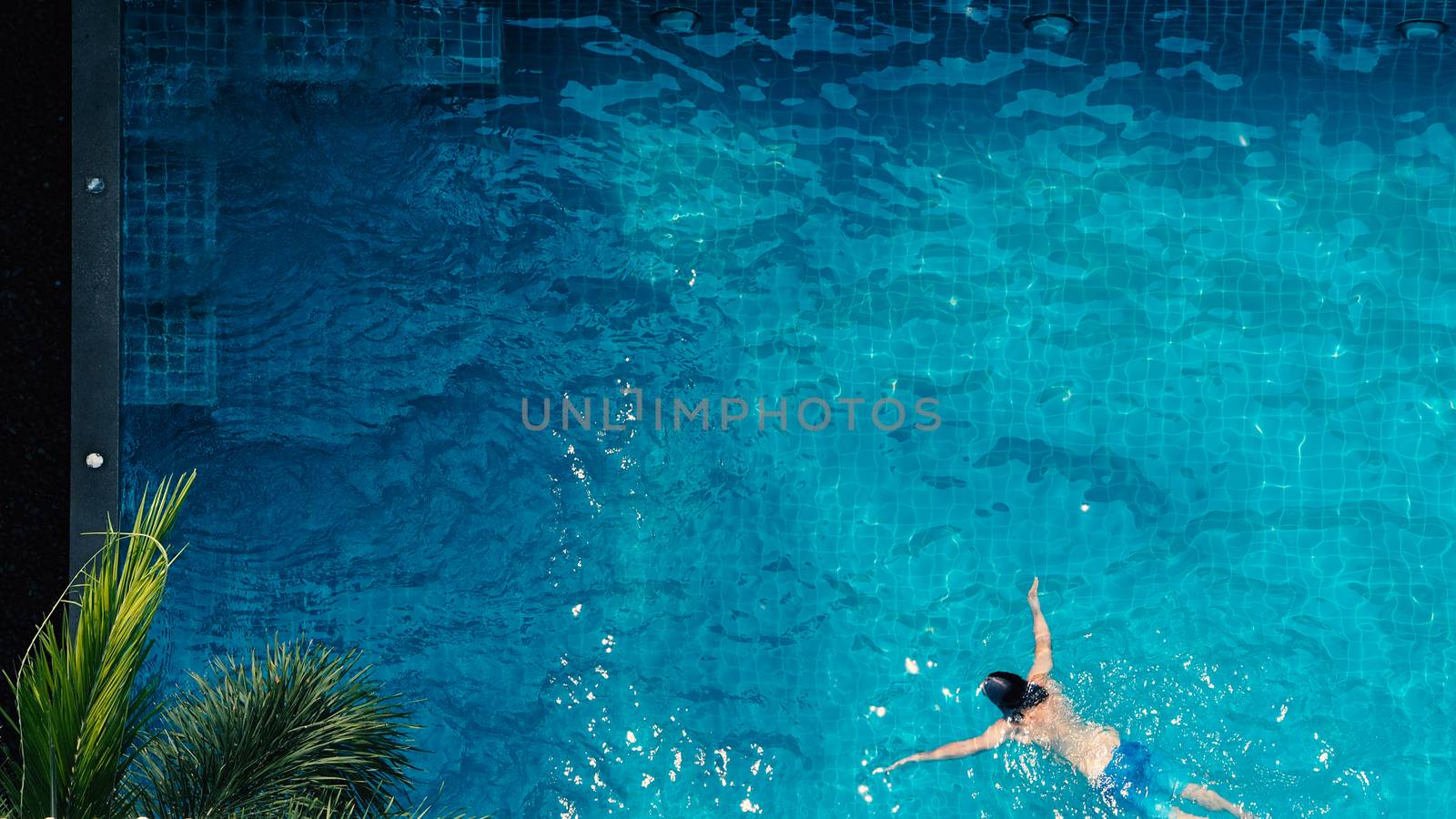 Swimming pool top view angle which young man relaxing and enjoying in pools of his hotel in summer season of Thailand at vacation day and have sun light and flares on blue water surface.