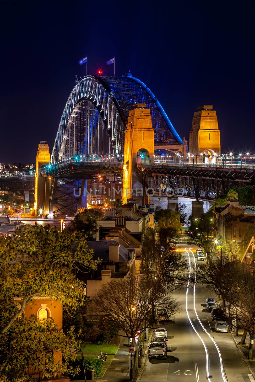 Views down the road towards Sydney Harbour Bridge, its arch lit with blue and the light reflecting in the harbour below.  A cars lights snake along the road.