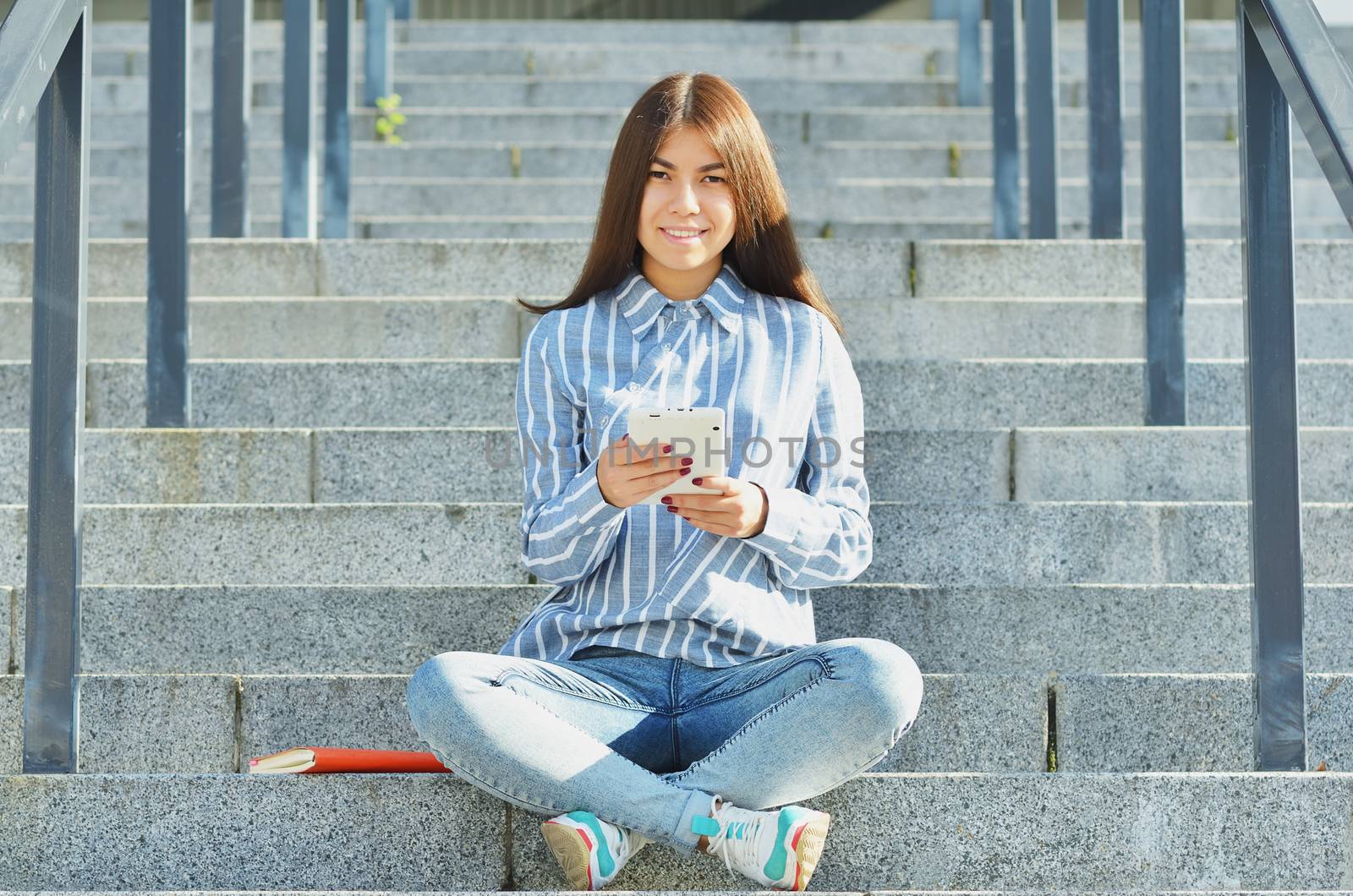 A young girl of Asian appearance, a student dressed in jeans and sneakers, holding a tablet and sitting on the stairs