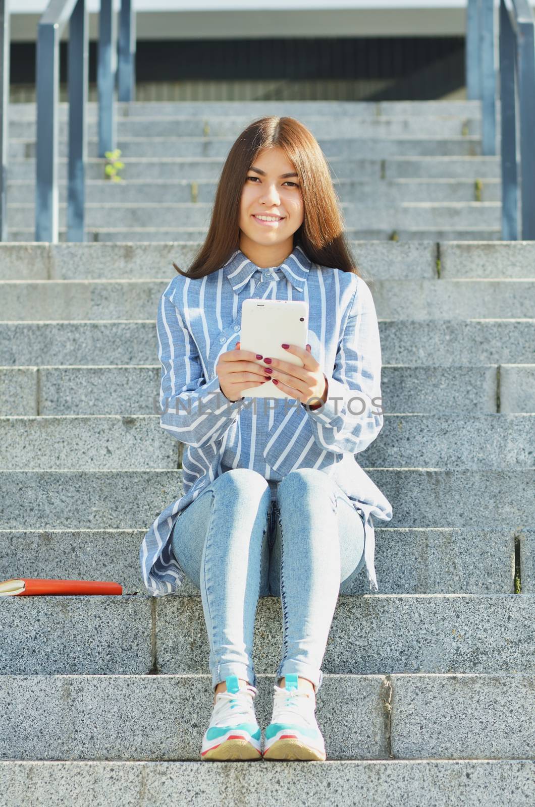 A young girl of Asian appearance, a student dressed in jeans and sneakers, holding a tablet and sitting on the stairs, and communicates by video