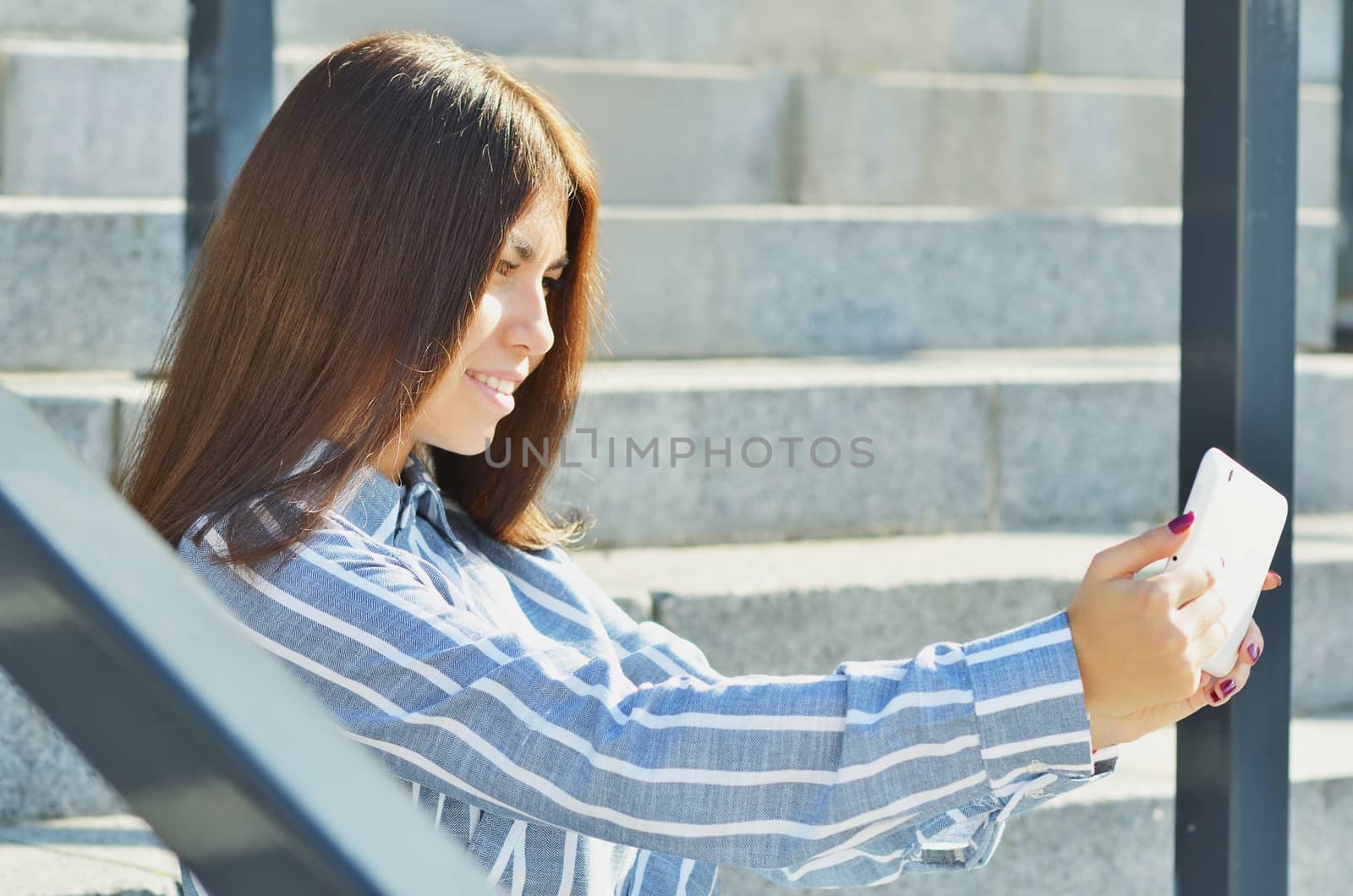 Young beautiful girl student Asian appearance, holding a tablet and talking on video, sitting on the steps in the street
