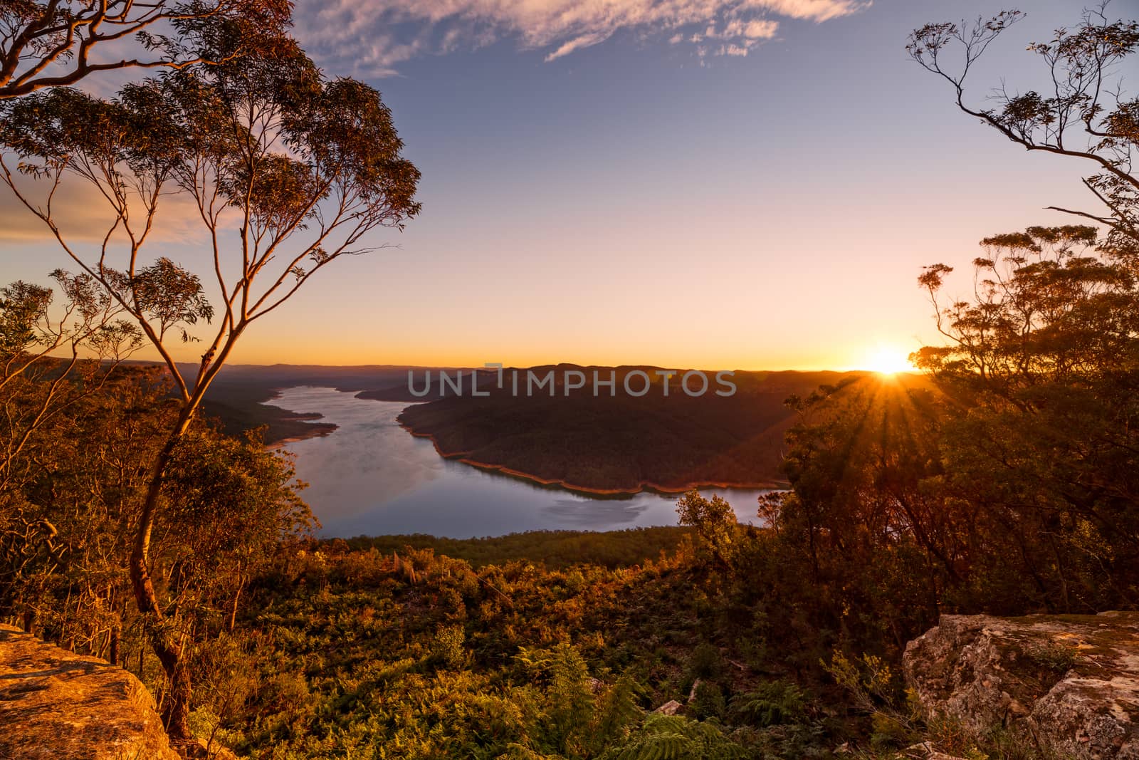 Sunset scenic vista from Nattai with Burragorang Lake in view. Rocky escarpment with large gum trees in the foreground as the sun sets on the horizon casting its last golden rays onto the landscape.