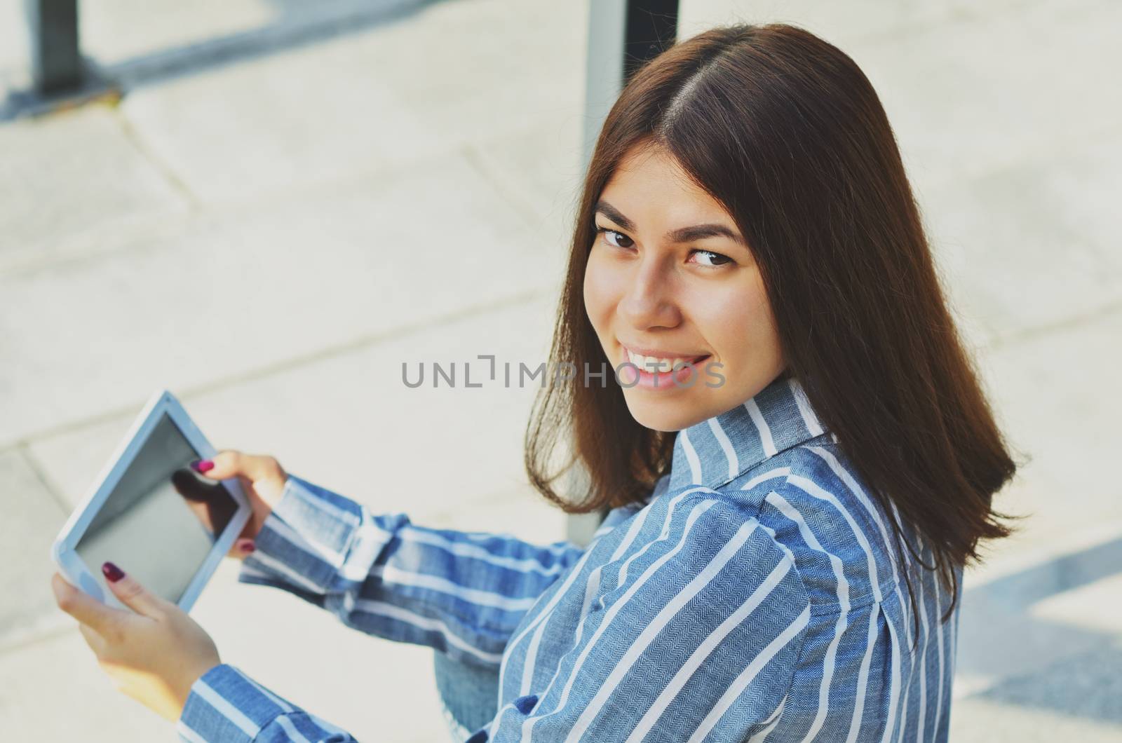 Young beautiful Asian woman smiling at the camera and holding a tablet in the street in Sunny weather