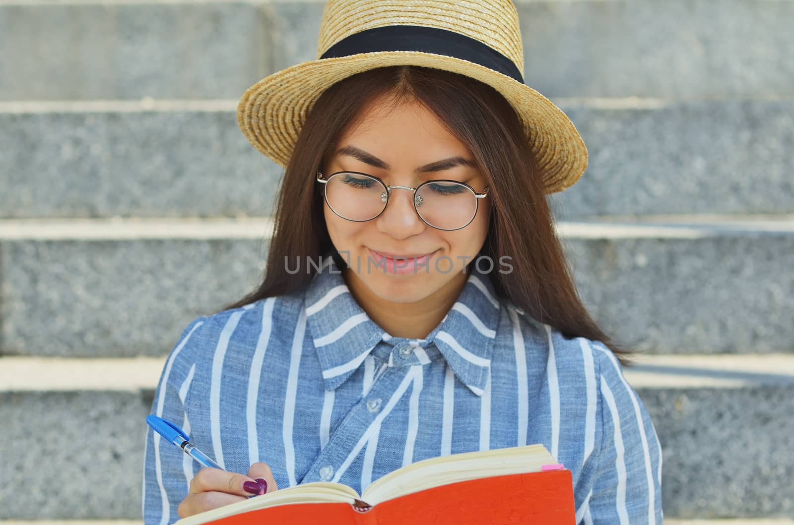 Portrait of a young Asian student in glasses with a hat dressed in a blue striped shirt that writes in a notebook task