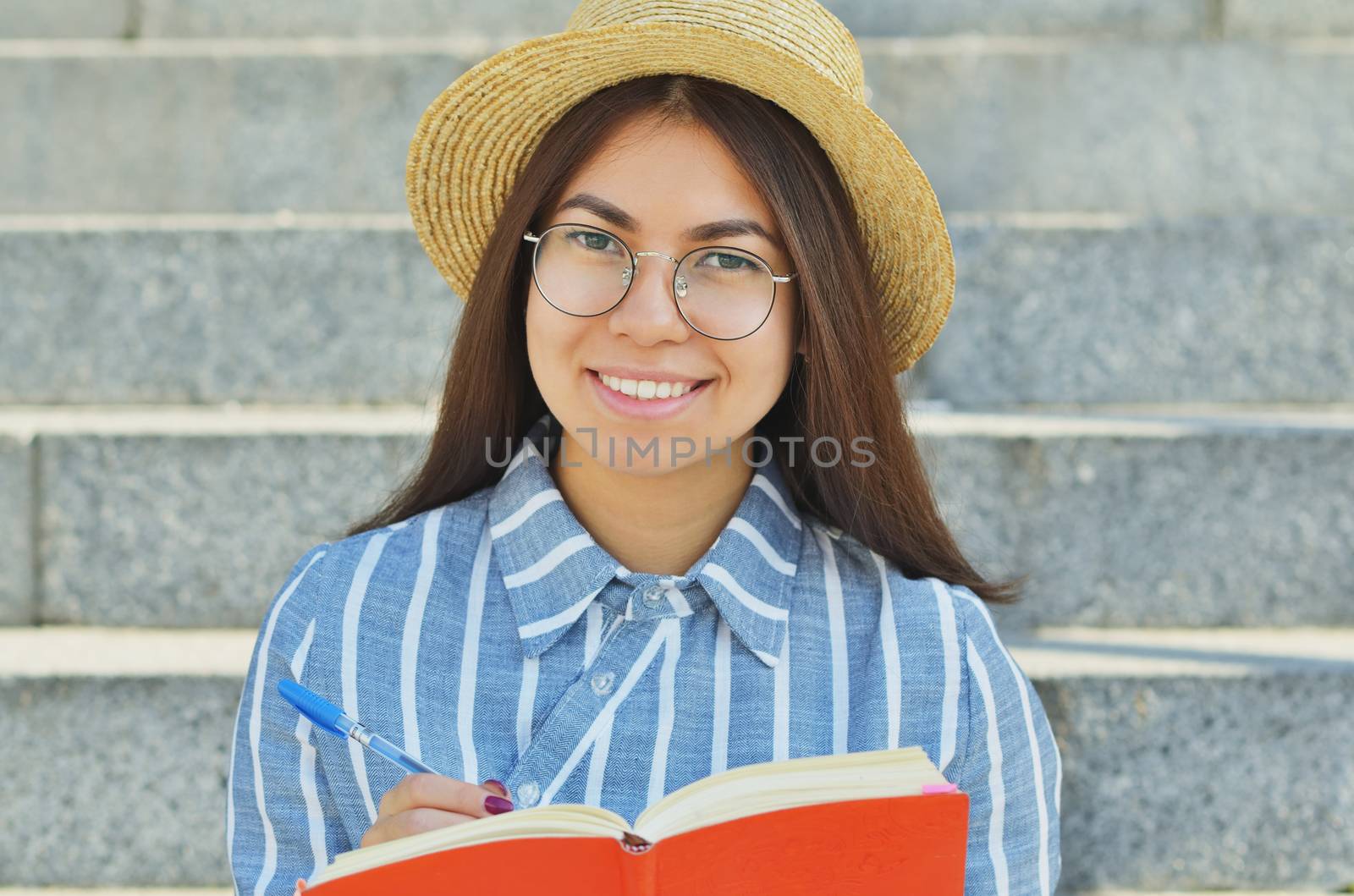 Portrait of a young Asian student in glasses with a hat dressed in a blue striped shirt who writes tasks in a notebook, and smiles at the camera