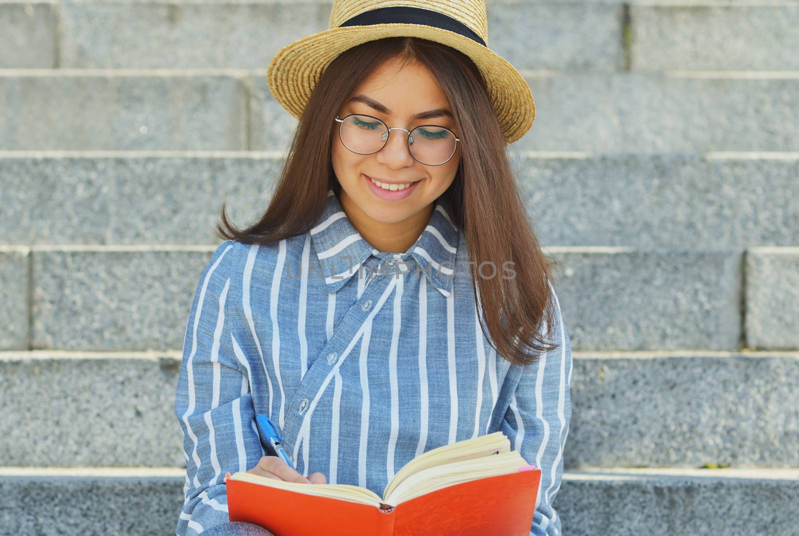 Portrait of a young Asian student in glasses with a hat dressed in a blue striped shirt who looks at the notebook writing tasks, smiling at the camera