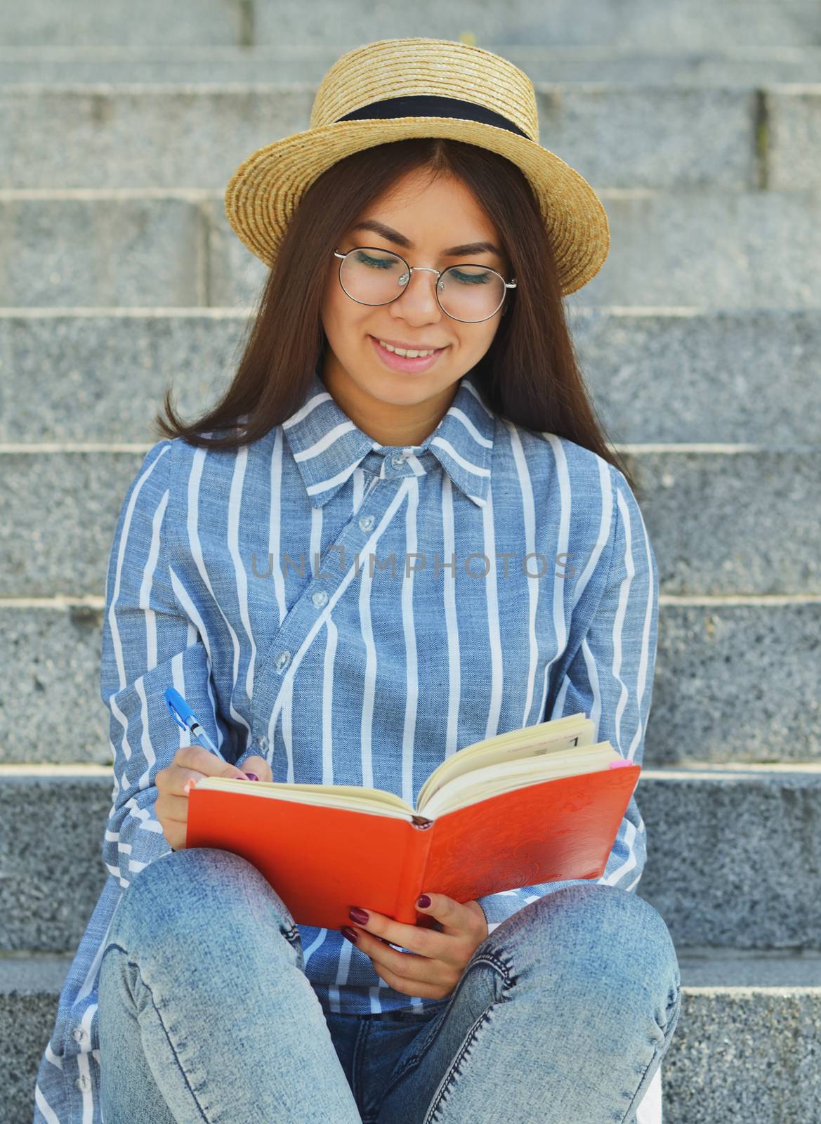 Portrait of a young Asian student in glasses with a hat dressed in a blue striped shirt that looks in a notebook writing tasks. Vertical photography