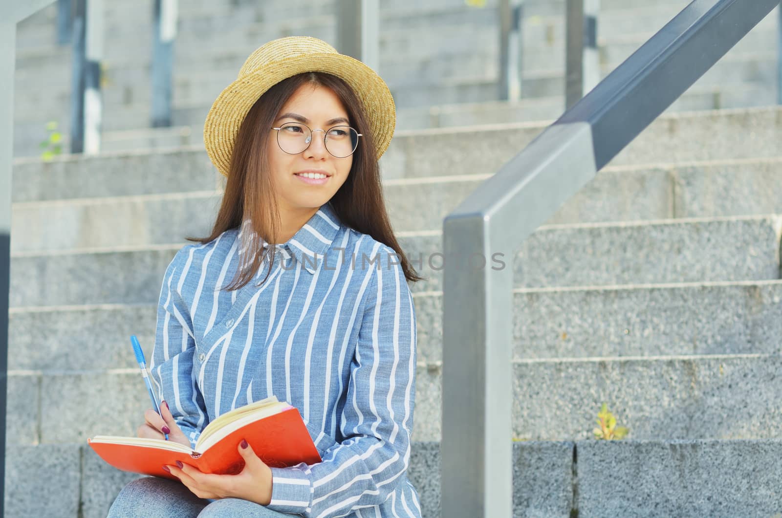 Portrait of a young Asian student in glasses with a hat dressed in a blue striped shirt that holds a notebook and writes tasks and looks into the distance