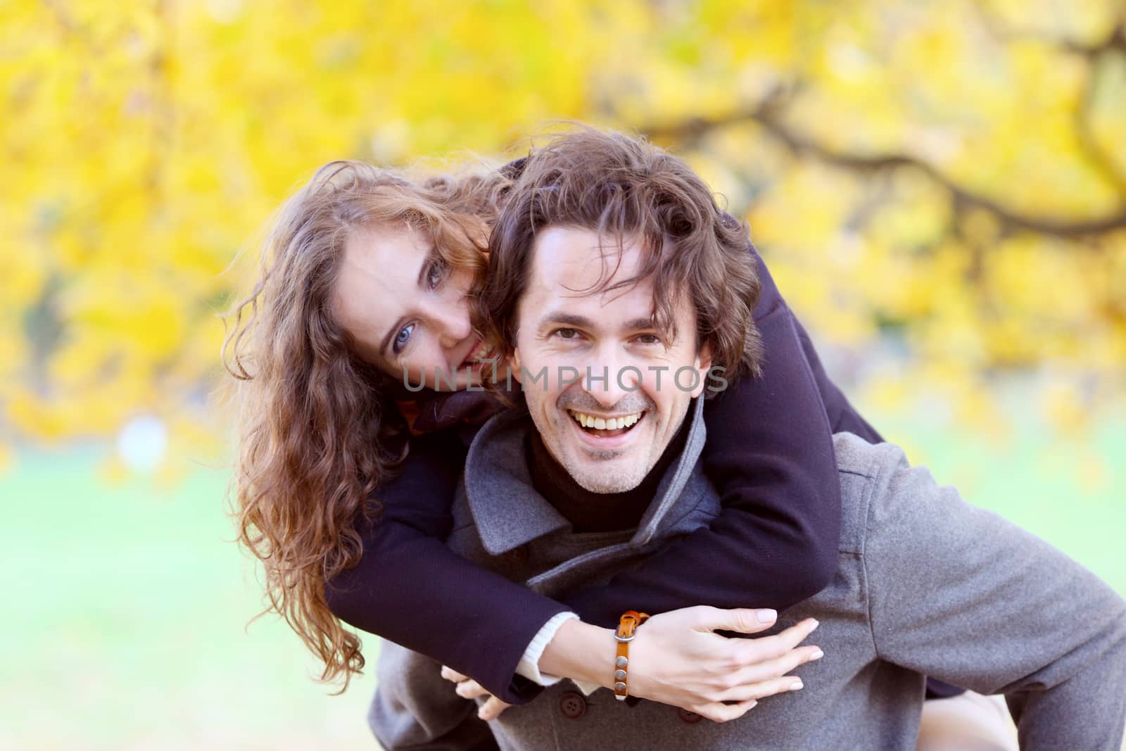 Smiling mid adult couple hug in autumn forest of yellow maple trees giving piggyback ride