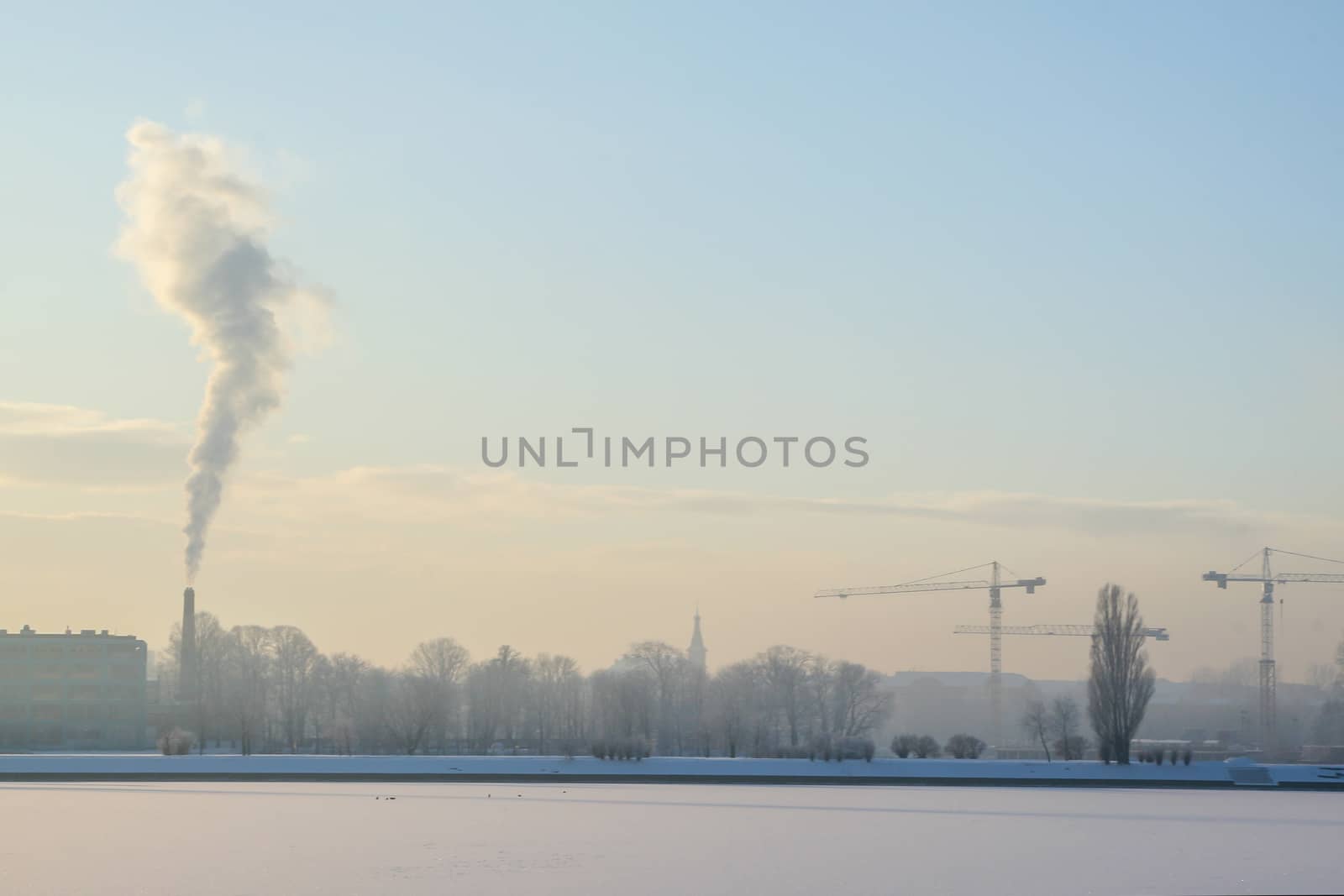 Riga view in winter; Riga, capital city of Latvia in winter time. View of Riga with frozen river Daugava and smoky chimney. City Riga with background of blue sky. 