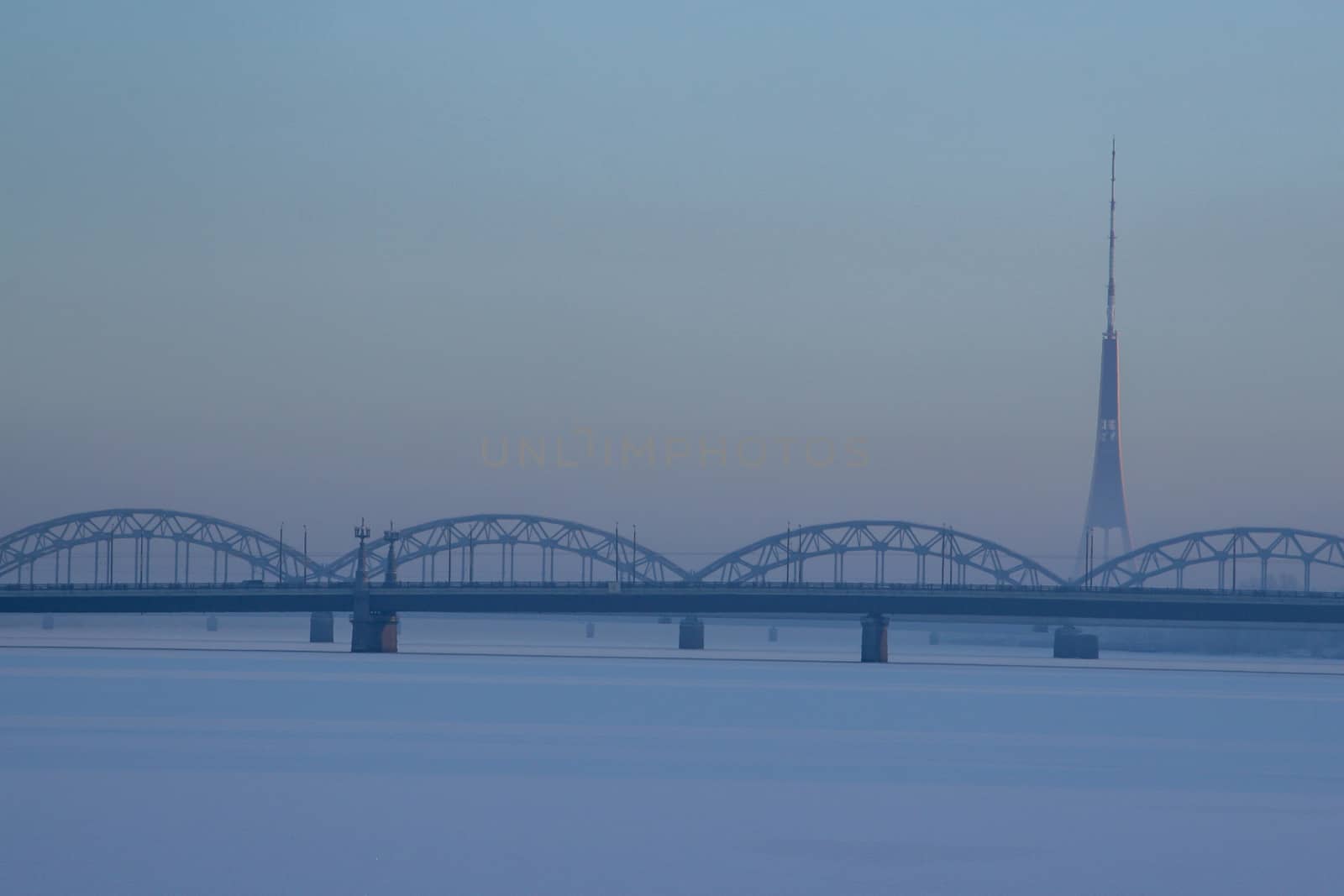 Railway bridge and television tower in winter. Railway bridge over the frozen river Daugava in Riga.TV tower and railway bridge on blue sky background in winter. Riga view in winter.