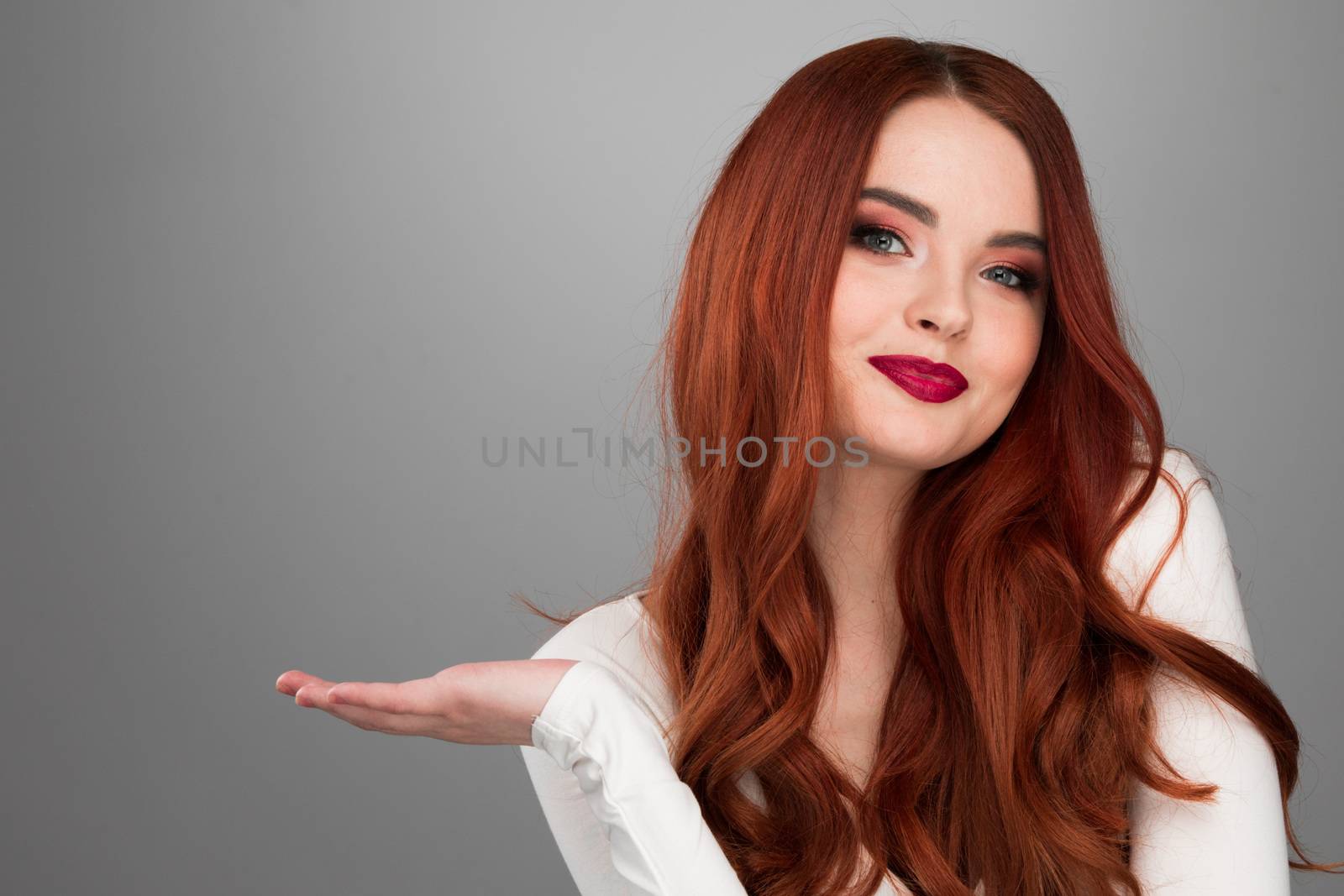Redhead girl showing copy space on her palm, looking at camera, standing over gray background