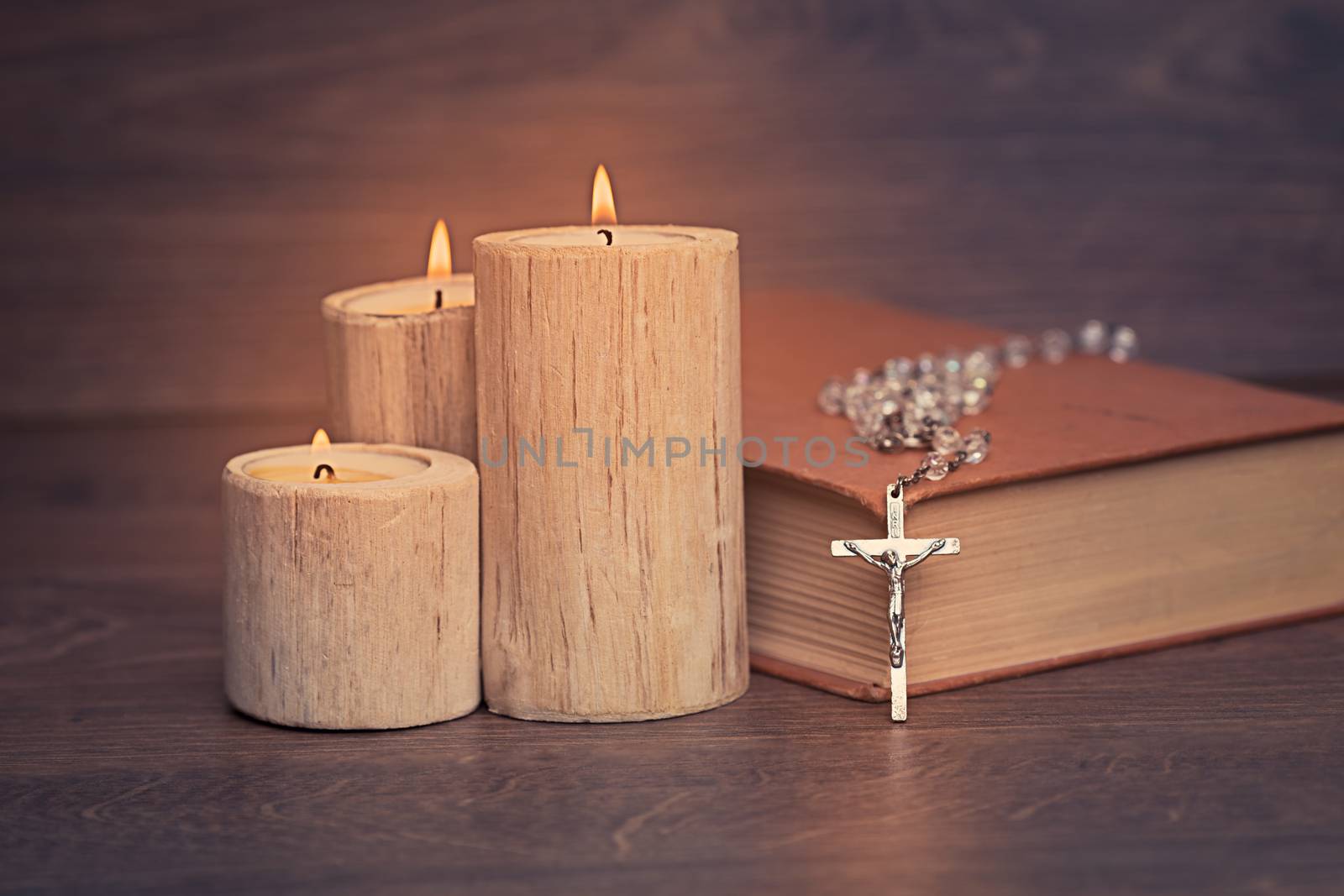 Silver rosary and crucifix resting on closed book near the candles on wooden table, religion school concept. Vintage style.