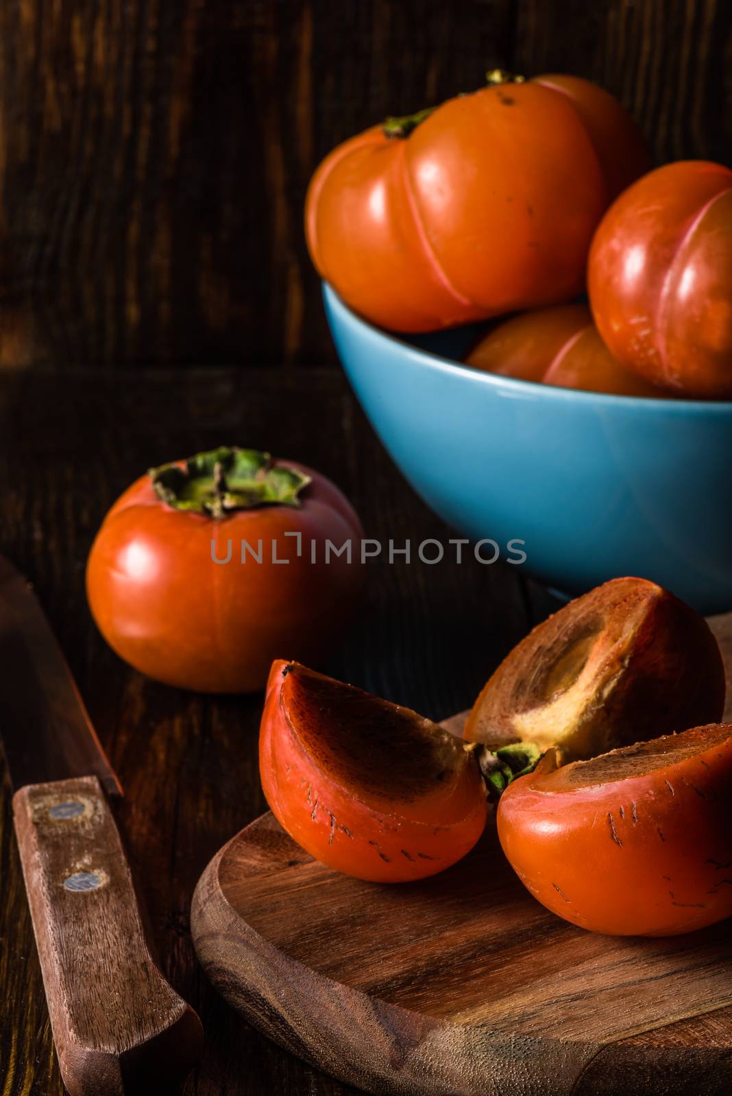 Sliced Persimmon for Three with Knife and Some Fruits in Blue Bowl on Background. Vertical.