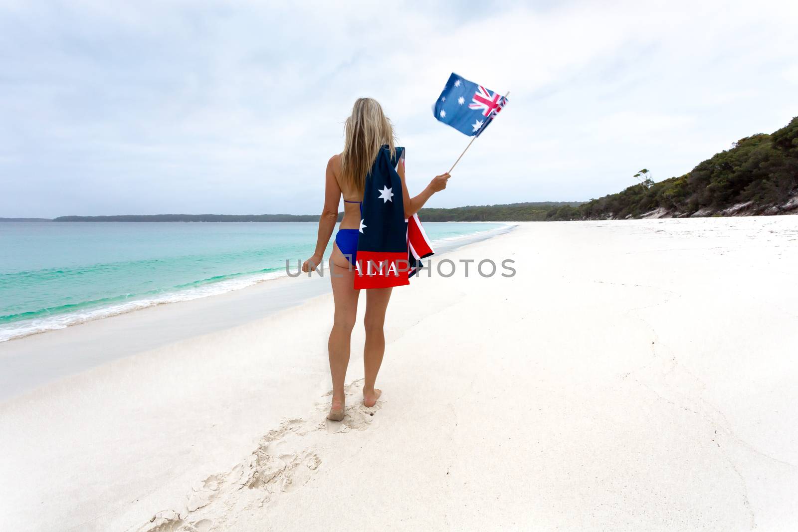 Aussie blonde woman walking along an idyllic beach wearing a bikini with patriotic Australian flag towel draped across one shoulder. She is also holding a small  flag.  Australian travel, Australia day, holiday, vacation.  Space for copy.