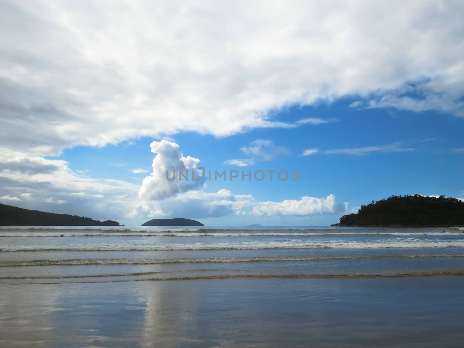 Beach with small waves in blue sky day and clouds, with islands on the horizon.