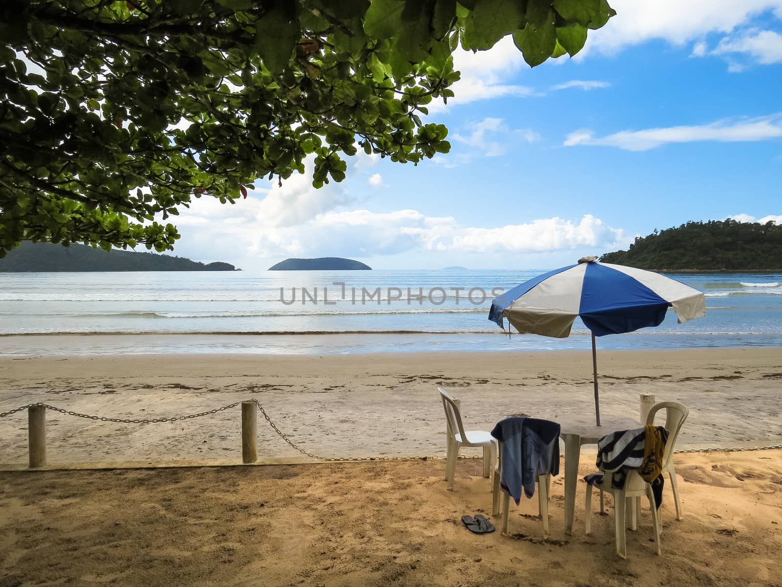 View of the beach, with chair and umbrella the shade of tree in the foreground, with coast and islands in the background, in Brazil.