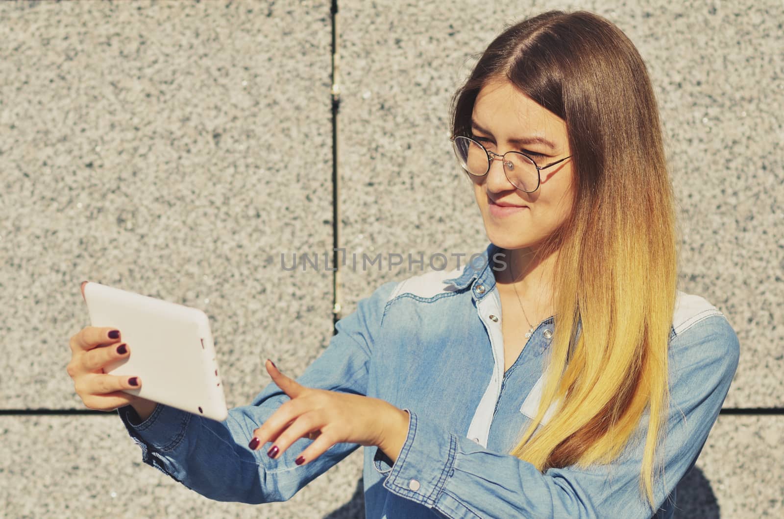 A young girl with glasses who is wearing a denim shirt, holding a tablet in her hand and clicks on the screen, horizontal photo