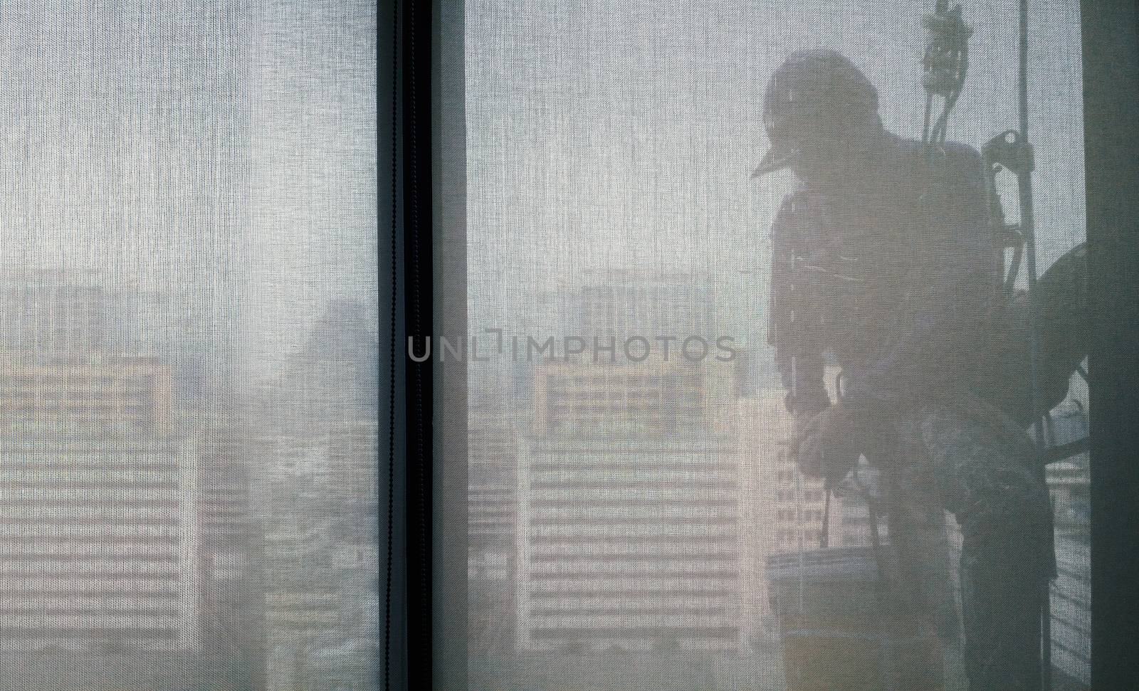 Silhouette images and shoot through transperent curtain effect from inside building which a man cleaning the window of high office building with his equipment such as wipe, sponge, bucket and high risk of danger