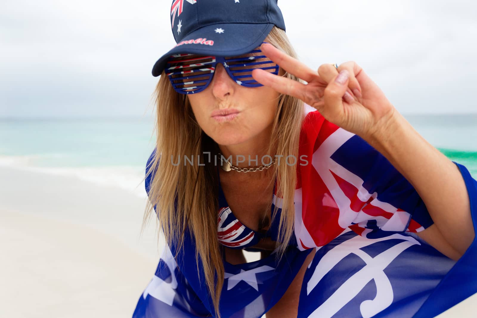 Proudly Australian woman lips pursed into a kiss and holding up two finger V good vibes.  She is proudly wearing the Australian flag on her clothes and accessories on a beach in Australia.