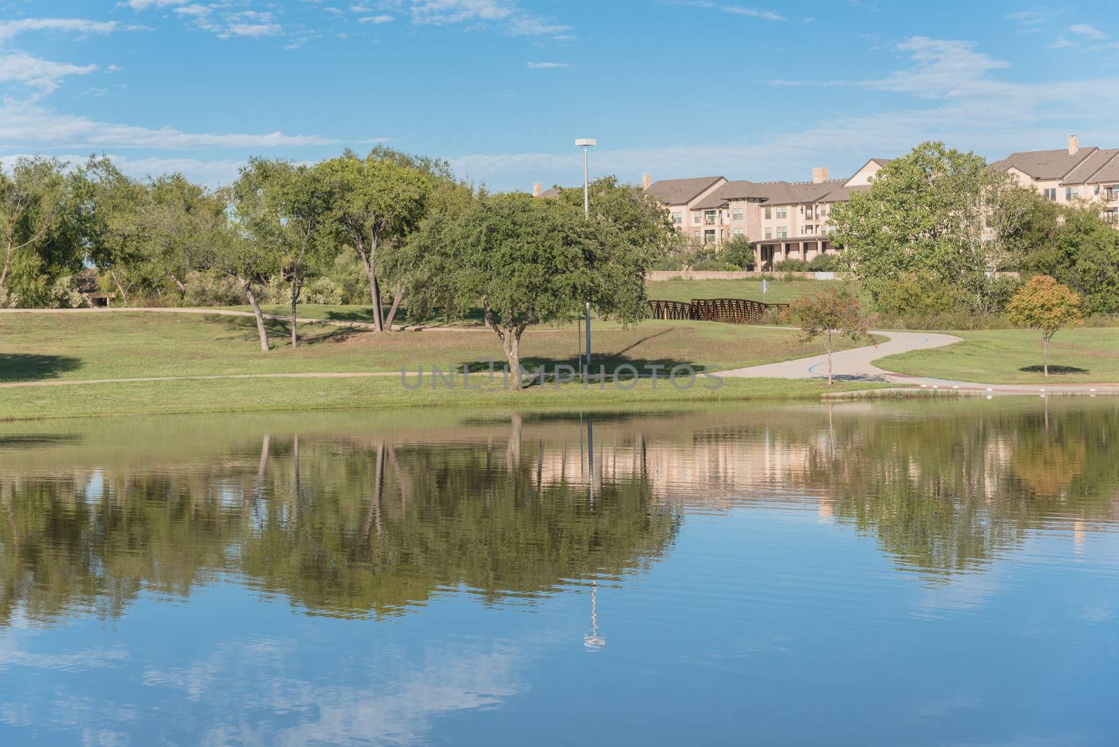 Multistory apartment complex near city park with pathway, lake and cloud sky reflection. Natural rental area near Dallas, Texas, USA