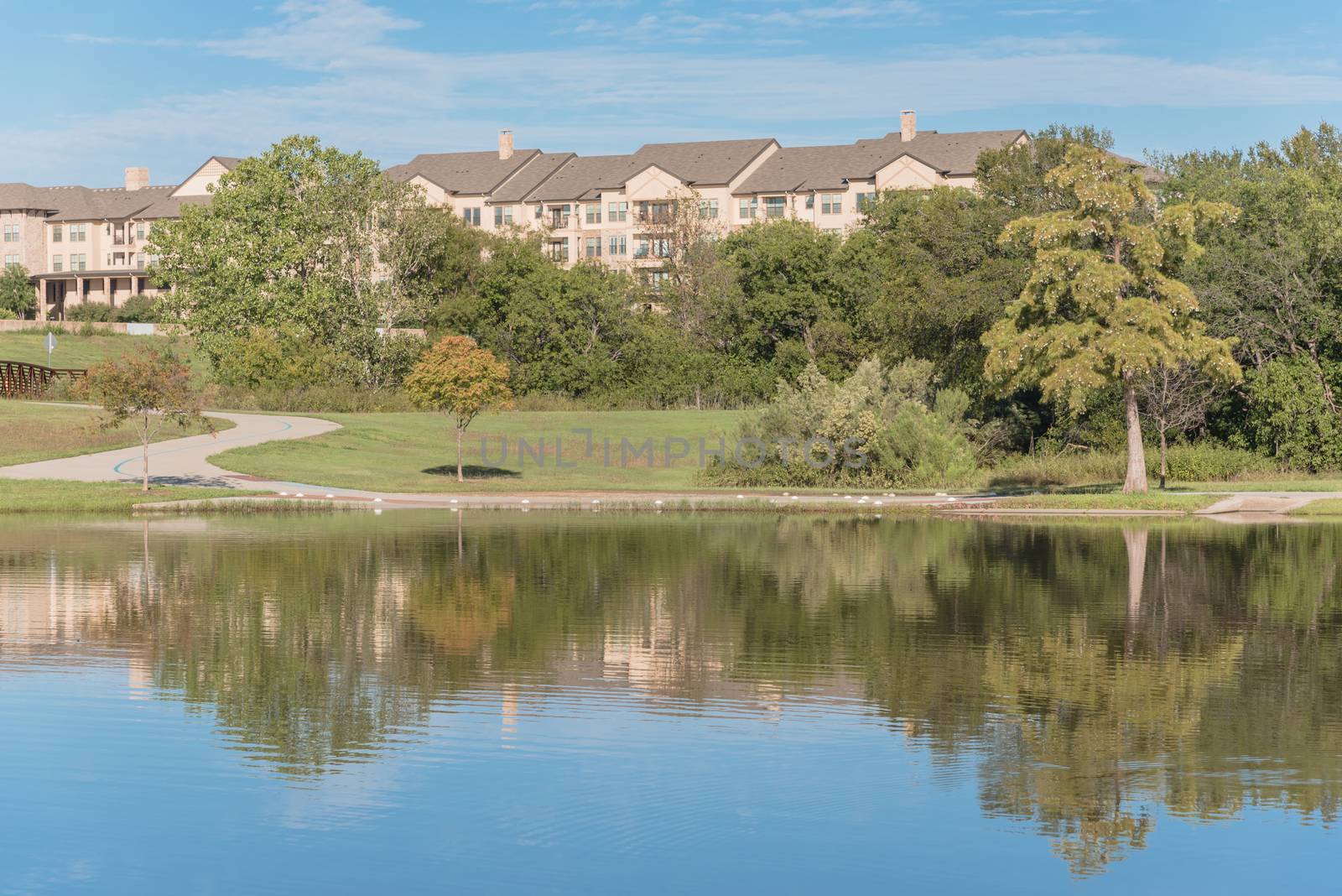 Multistory apartment complex near city park with pathway, lake and cloud sky reflection. Natural rental area near Dallas, Texas, USA