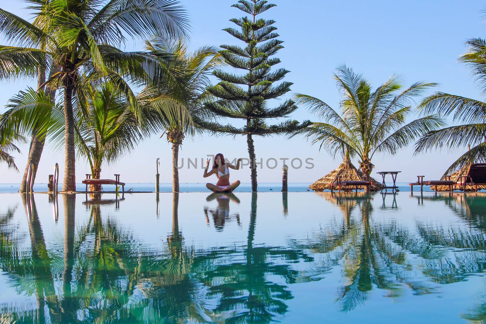 Beautiful slim woman sitting by the pool at tropical resort blue sea on background and palms around