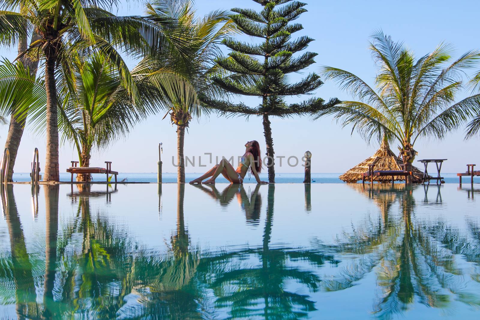 Beautiful slim woman sitting by the pool at tropical resort blue sea on background and palms around
