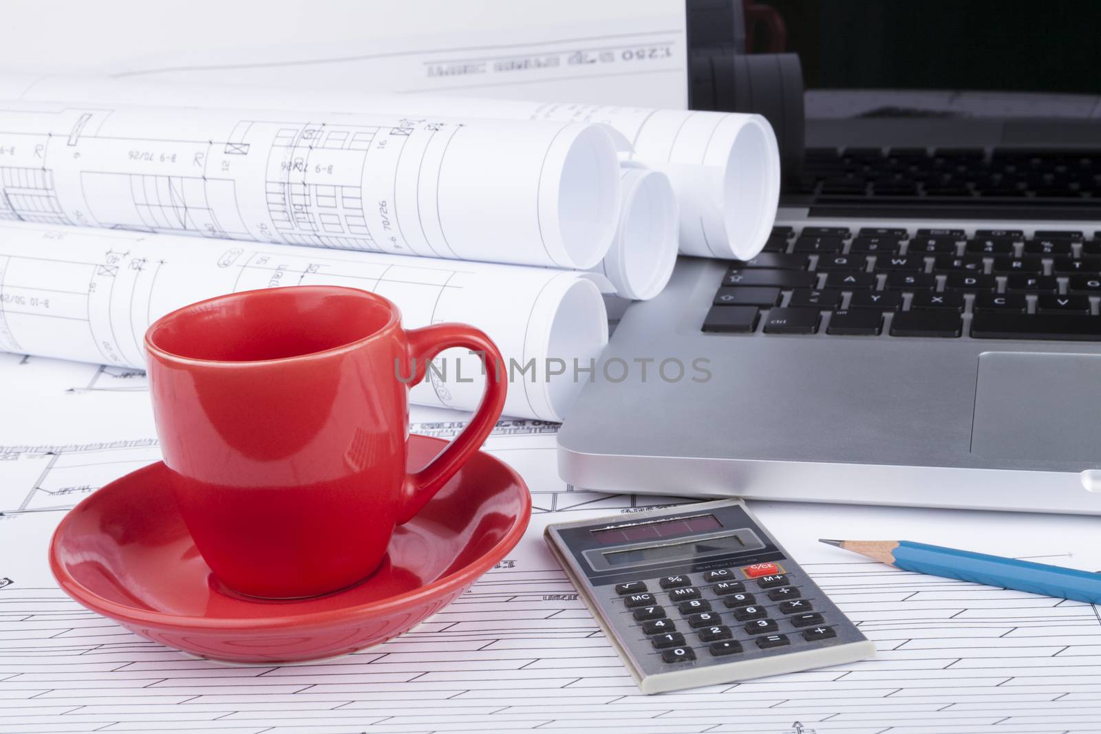 Architect blueprints with a calculator and a silver laptop with blue pencil and red cup of coffee